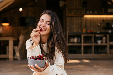 Attractive young caucasian girl enjoys eating ripe cherries sitting in campsite. Brunette with closed eyes relaxes. Sincere emotions lifestyle concept