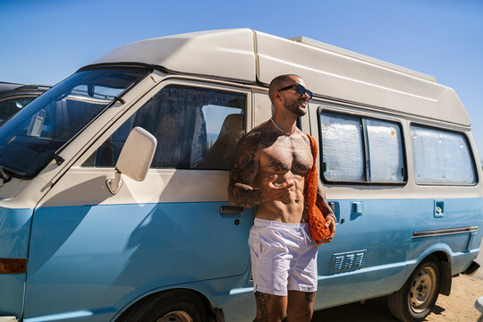 Chico joven tatuado y musculoso posando y sonriendo delante de una furgoneta camper vintage