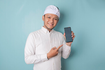 Excited Balinese man wearing traditional headband or udeng and white shirt pointing at the copy space on his smartphone, isolated by blue background