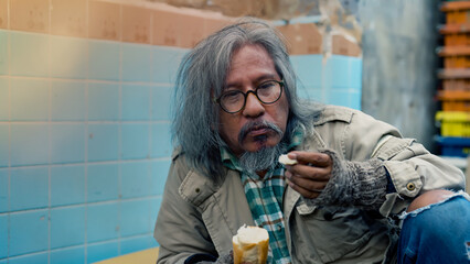 The head of a long-haired, homeless man sits at night eating bread donated by passers-by in the...