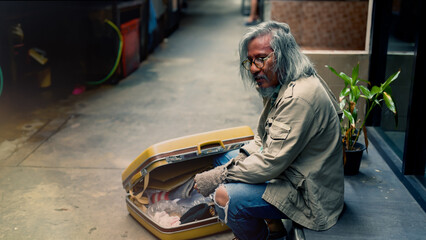 A homeless old Asian man with his big yellow bag is sitting on the ground in a narrow alley and opening his bag to find something to eat. Homeless people who do not have home to sleep on streets