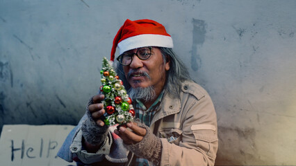 Old homeless Asian man wearing a red Christmas hat holding a small Christmas tree is showing signs...
