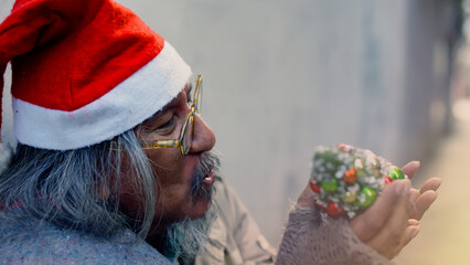 An old homeless Asian man wearing a red Christmas hat holding a small Christmas tree is showing...