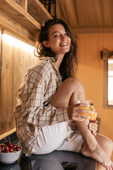 Pretty young caucasian woman with juice smiling looking at camera sitting on tabletop camping. Brunette wears light shirt and shorts. Lifestyle, different emotions, leisure concept.