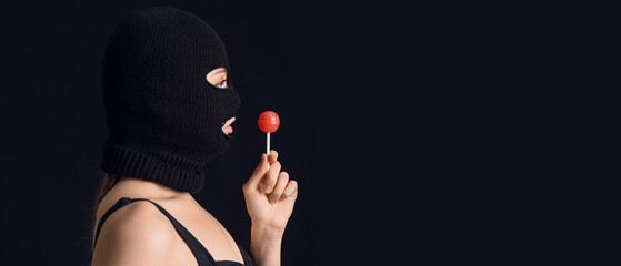 Young woman in balaclava and with lollipop on black background with space for text