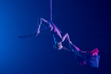 Female circus gymnast hanging upside down on aerial silk on black background with blue backlight....