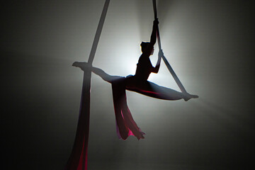 Dark silhouette of a young woman equilibrist demonstrating twine, hovering on an airy silk at...