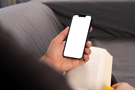 Holding Mobile Phone, Over Shoulder View Of Man Hand Holding Mobile Phone.  Blank White Screen For Smartphone Mock Up. Caucasian Male Relaxing On The Couch, Reading Book And Looking To Phone Display.