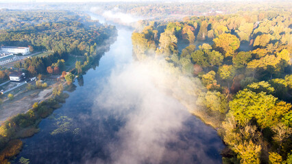 Aerial drone view flying over the river with calm reflective water surface and white fog mist and forest trees on the banks of the river on a sunny early autumn morning. Beautiful natural background.