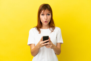 Redhead girl isolated on yellow background looking at the camera while using the mobile with surprised expression