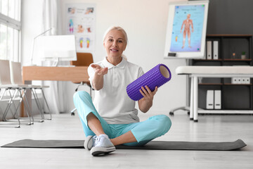 Fototapeta na wymiar Mature physiotherapist with foam roller sitting on mat in rehabilitation center
