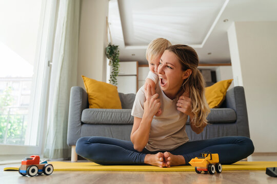Funny picture of cheerful mother sitting in butterfly position on mat practicing yoga piggybacking her cute baby boy in living-room with gray couch on background. Healthy motherhood and wellness
