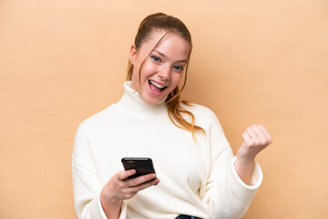 Young caucasian woman isolated on beige background with phone in victory position