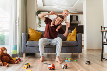 Happy father carrying his laughing little blond son on neck and sitting on couch in living-room...