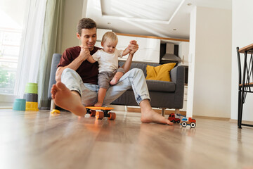 Young father playing with his little baby boy in living-room, holding his hands while kid trying to stand on skateboard. Active games at home. Carefree childhood, happy fatherhood. Male babysitter