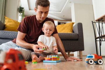 Father assisting his baby boy playing sorter sitting on floor in living-room with sofa on background, surrounded with car toys, waiting mom to come home from work. Education toys, child development