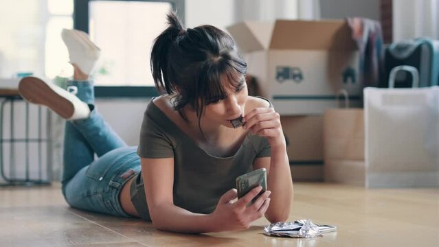 Video of  young beautiful woman with mobile phone while eating a piece of chocolate lying on the floor in her new house.