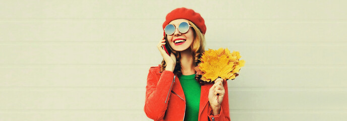 Autumn portrait of happy smiling young woman with yellow maple leaves calling on phone wearing french beret over gray background