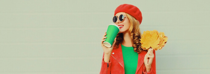 Autumn portrait of happy smiling young woman with yellow maple leaves and cup of coffee wearing red french beret on gray background