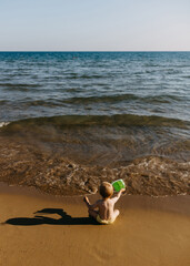 Fototapeta na wymiar Little child sitting alone on a seashore playing with a green bucket