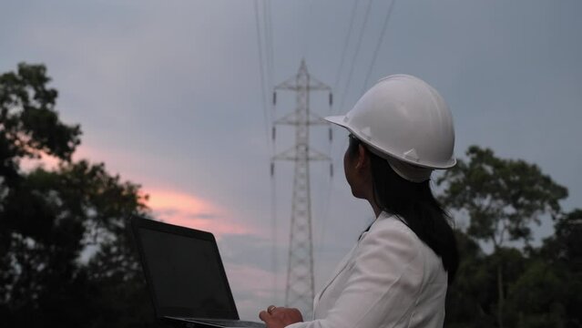 Asian female electrical engineer working on laptop near high voltage pole, inspecting power grid. Wind Turbine Engineer for Electric Power Generation. renewable energy concept