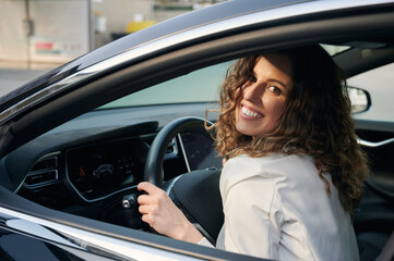 Fototapeta na wymiar Portrait of business woman with sincere smile on face who loving her car. Curly joyful girl sitting in cab of her auto, holding hands behind the wheel.