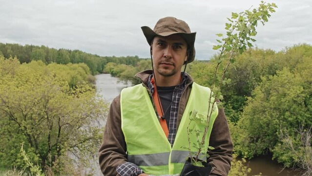 Medium of male gardener in work clothing and reflective vest with seedlings, standing in foreground of river in forest on cloudy day