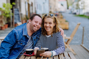 Portrait of happy young man with Down syndrome with his mother sitting at cafe outdoors and talking.