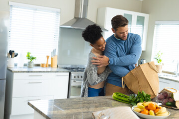 Image of happy diverse couple embracing and unpacking groceries in kitchen