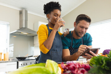 Image of happy diverse couple preparing meal together, drinking wine and using tablet