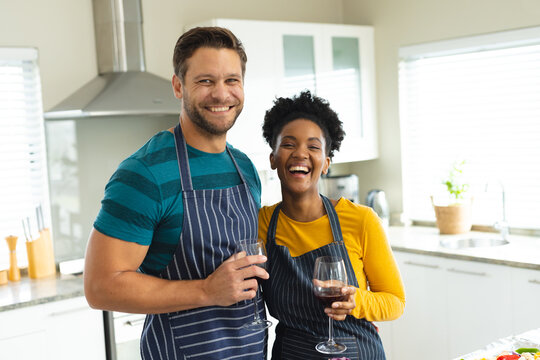 Image of happy diverse couple in aprons with wine in kitchen
