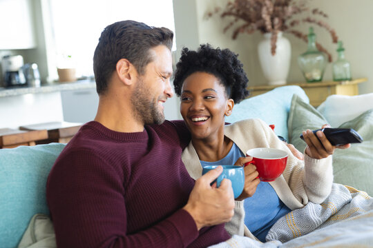 Image of happy diverse couple sitting on sofa with coffee and watching tv