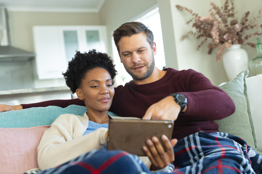 Image of diverse couple using tablet and sitting on sofa