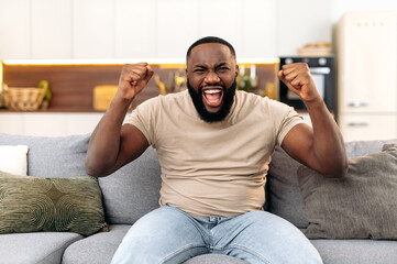 Winning concept. Amazed cheerful young african american man, sitting on the sofa in living room, excited for success with arms raised, celebrating victory, rejoices at a goal, smile happily, screaming