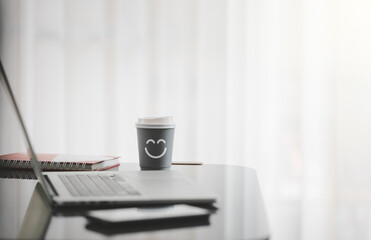 smiling face on paper cup of hot coffee with laptop on desk besides window