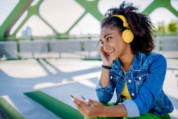 Multiracial teenage girl with backpack, headphones and smartphone resting after school in city bridge, listening to music.