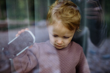 Little child standing alone with car toy behind the window, photo trough glass.
