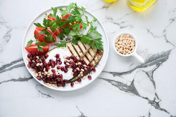 Grilled eggplant with pomegranate, yogurt, tomatoes and parsley, flat lay on a black and white marble background, horizontal shot with space