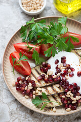 Grilled aubergine slices with pomegranate, pine nuts, white yogurt, tomatoes and parsley on a beige plate, close-up, vertical shot