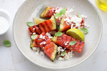 Beige plate with grilled watermelon batons, grated cheese and lime wedges, middle close-up, selective focus, horizontal shot
