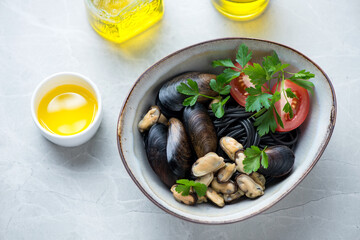 Bowl of mussels with black spaghetti, tomatoes and parsley, elevated view on a light-grey stone background, horizontal shot