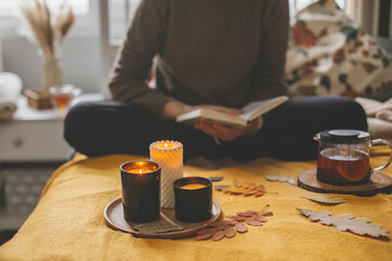 Woman reading a book on bed, drink tea. Burning candle and seasonal autumn home decor. Morning routine, home lifestyle concept.