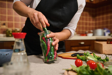 Focus on the sterilized glass jar and the chef's hands filling it with red and green hot chili while preparing fermented vegetables in the home kitchen. Canning. Pickling. Concept of food preservation