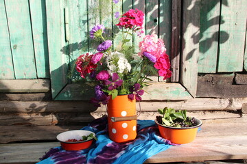 Rustic still life with garden flowers in can, aronia berries and strawberry in bowl on textile cover on old wooden background in sun light