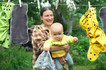 Mother with little baby hang reusable diapers to dry on clothesline in backyard garden. Modern eco friendly cloth nappy for infant child hygiene. Sustainable lifestyle, zero waste concept.