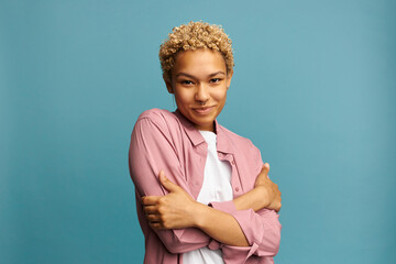 Pretty shy african america young woman with blond curls posing on blue background with crossed...