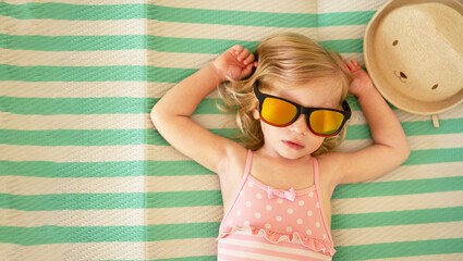 Blonde caucasian toddler, baby girl lying on striped towel on beach with sunglasses, swimsuit sunbathing,taking sun.Kid,white skin tan concept, child health care, copyspace.
