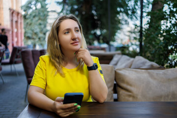 a pensive girl in a yellow dress sits at a cafe table and holds a phone in her hand