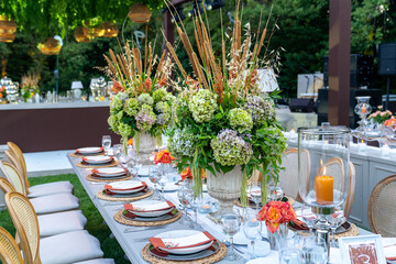 Beautiful decoration of a wedding banquet with a meal in the green garden in white colors. Very stylish setting with tropical flowers in a vase, glass candlestick set, napkins, plates, glasses cutlery