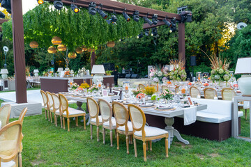 Beautiful decoration of a wedding banquet with a meal in the green garden in white colors. Very stylish setting with tropical flowers in a vase, glass candlestick set, napkins, plates, glasses cutlery